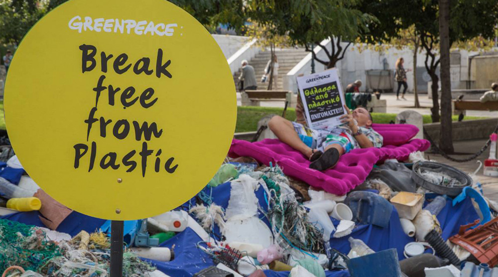 A man on the background is lying down on a purple bed on top of a pile of garbage collected during the brand audit 2018 in Greece. On the foreground is a yellow sign with the logos of Greenpeace and Break Free From Plastic