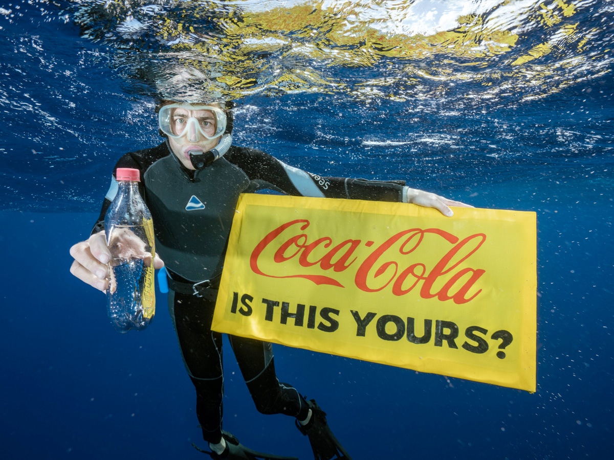 A diver underneath the ocean holding a coca-cola plastic bottle while also holding a sign saying "Coca-cola, is this yours"