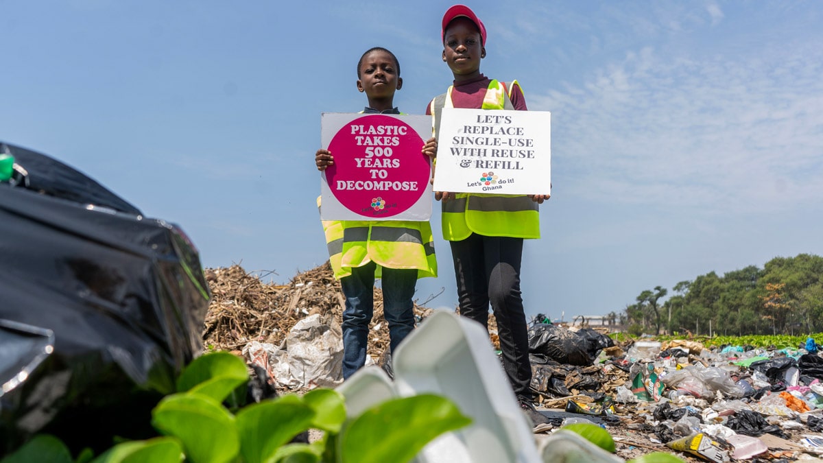 2 children who are volunteers from Let's Do it Ghana! hold up signs that say "Plastic takes 500 years to decompose" and "Let's replace single-use with reuse & refill". Photo by Kate Adobaya